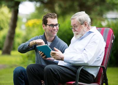 Älterer Mann sitzt im Park auf einem Stuhl, während er von einem jungen Mann etwas auf einem Tablet erklärt bekommt. 